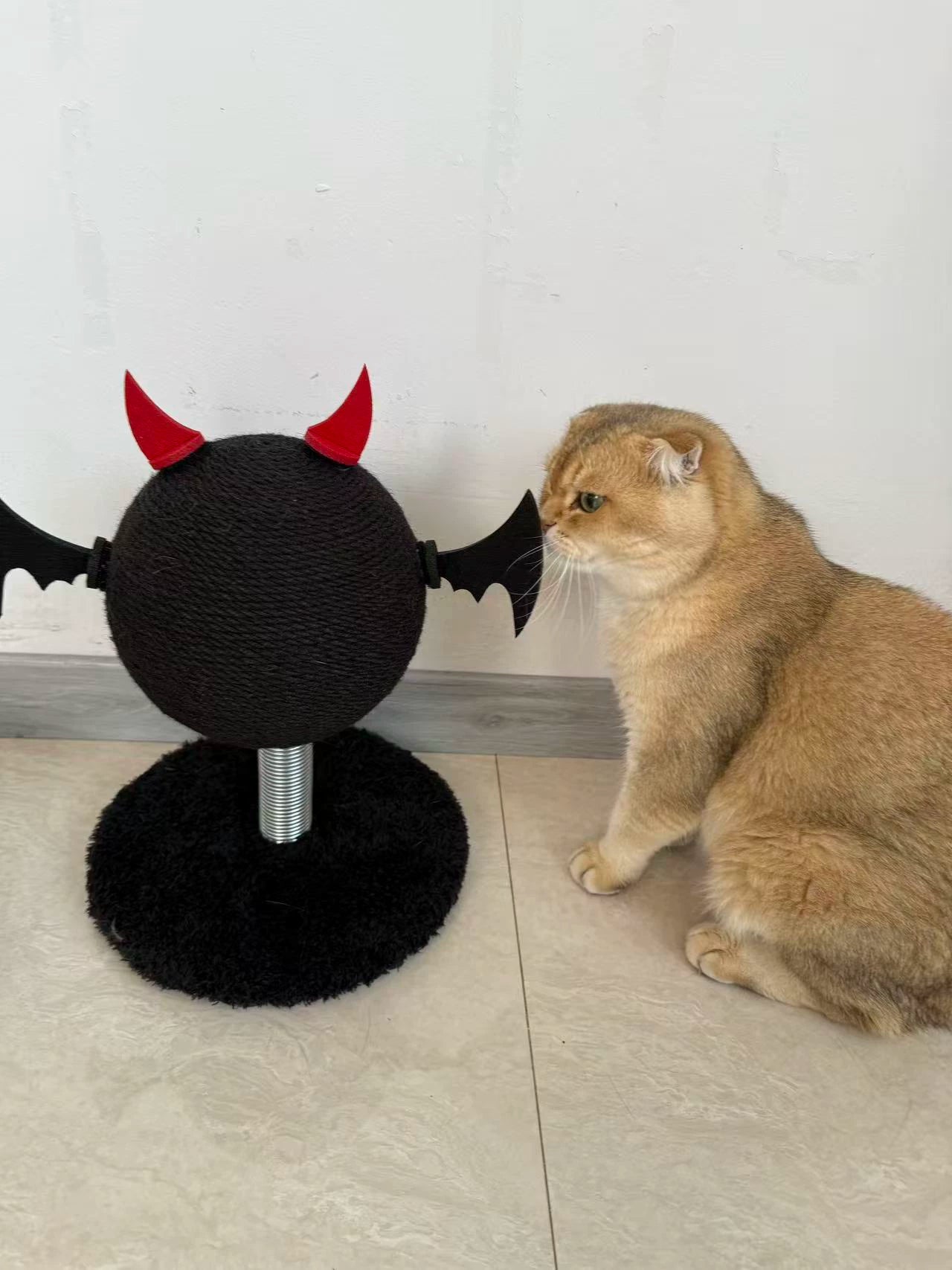 Cat sitting next to a black cat toy with red devil horns on a light-colored floor.